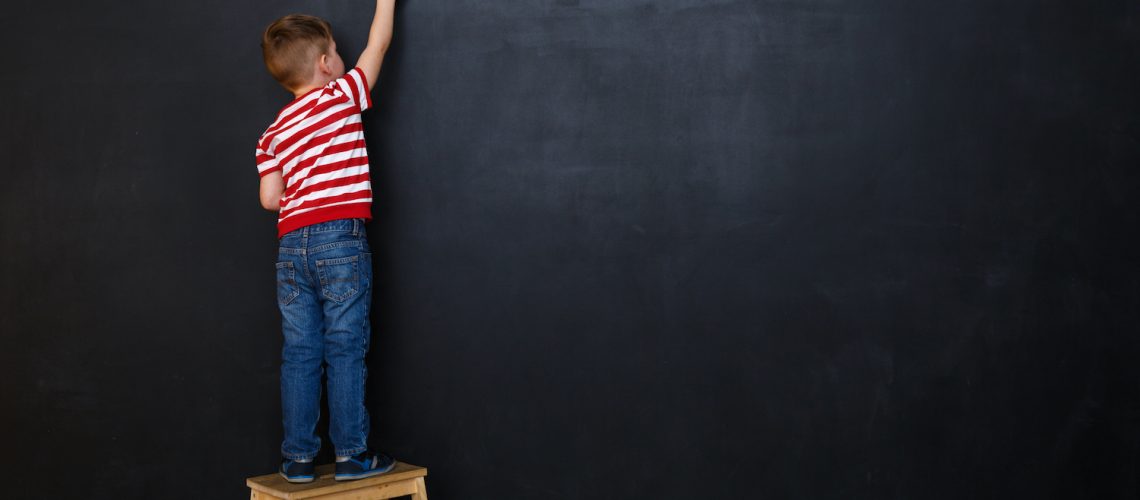 Back view of a little boy standing on ladder and writing with chalk on the backboard in school class